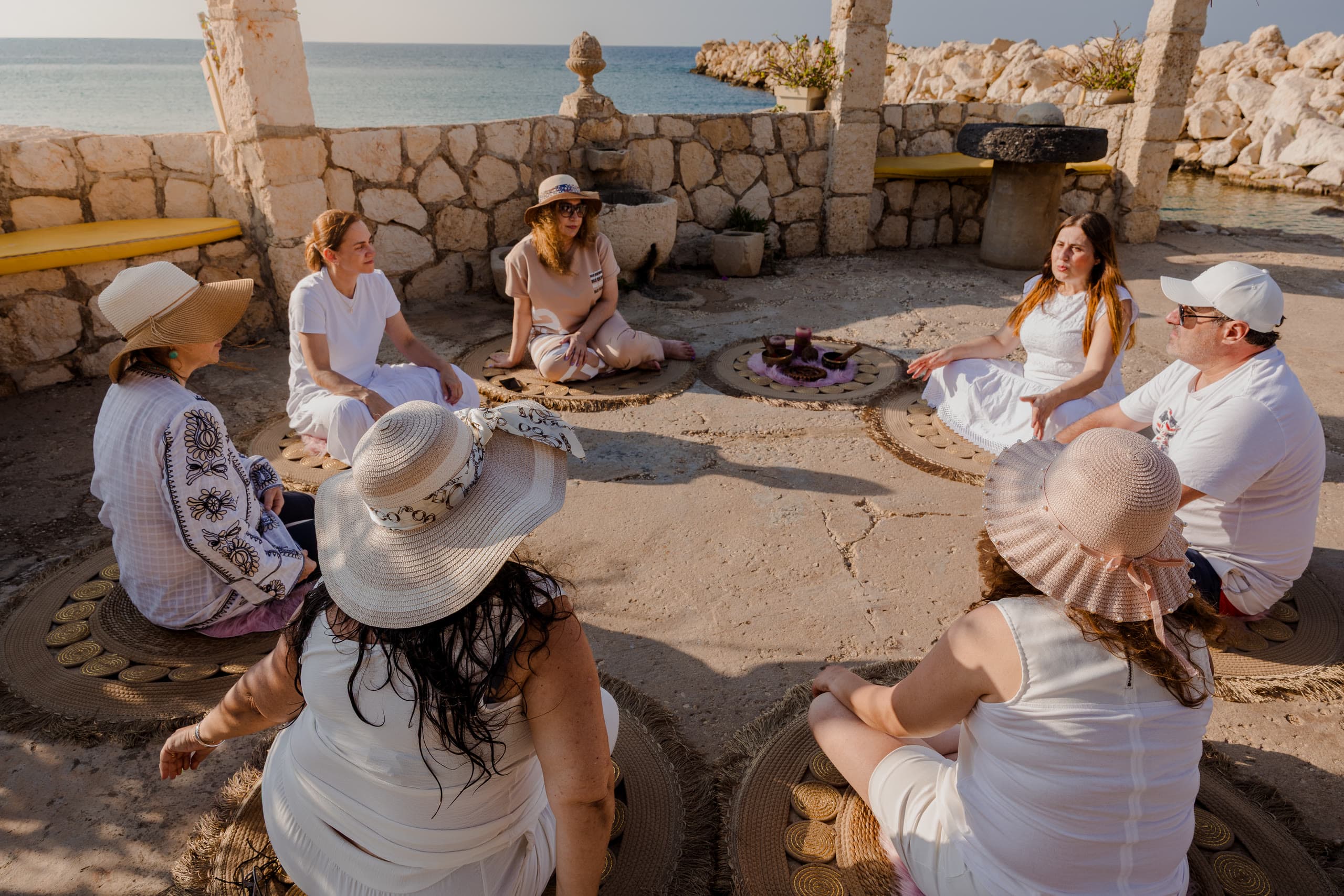 A circle of people seated on woven mats around a small altar at a seaside terrace in Lebanon, the sea behind them